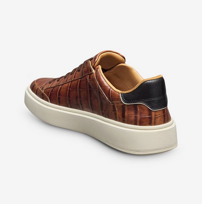 The Allen Edmonds Oliver Sneaker features brown premium leather with a crocodile texture, a white sole, and black heel detailing—shown from the back left angle—offering comfort and style for travel.