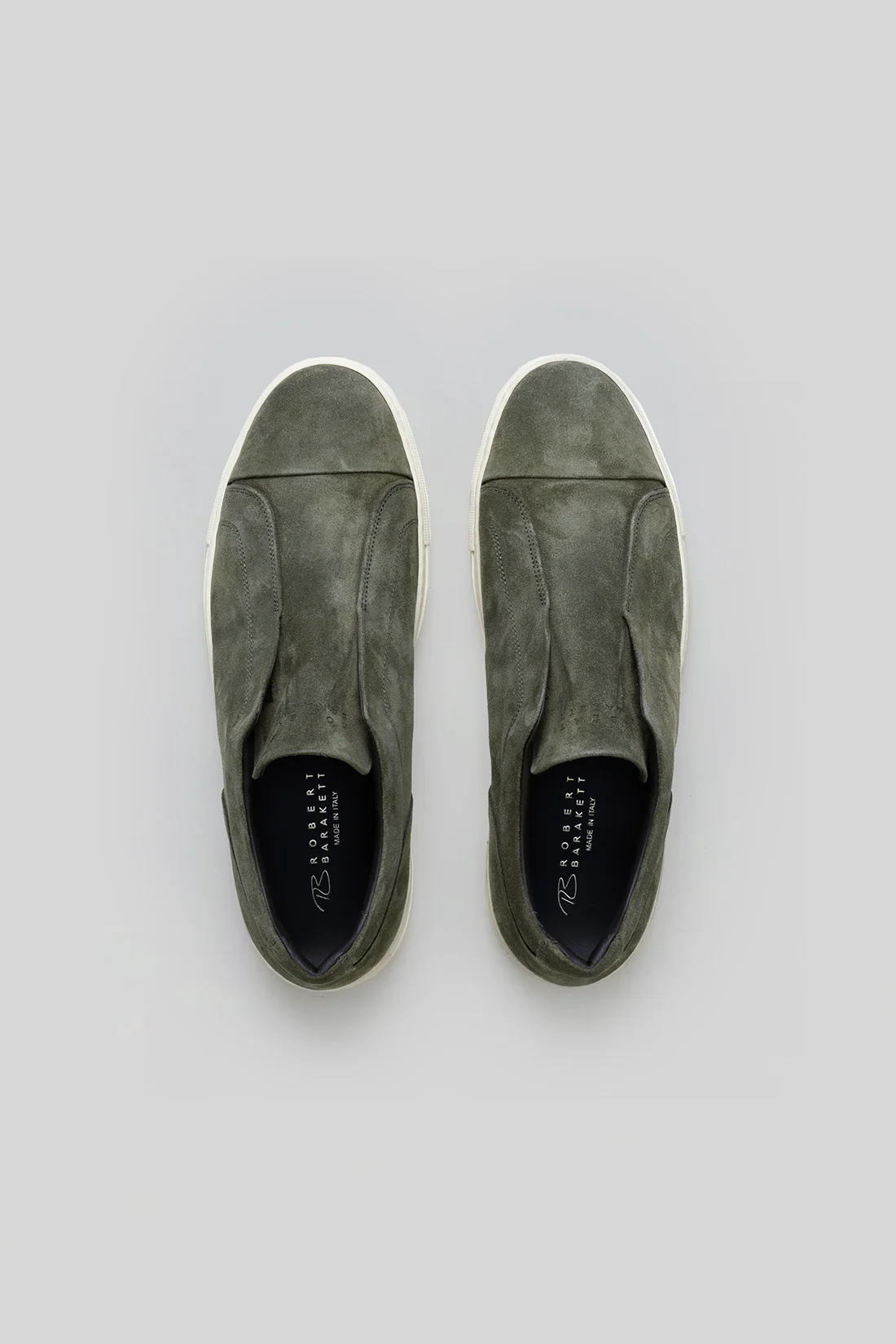 The Robert Barakett Catro Suede Sneaker by Robert Barakett is an olive green Italian suede slip-on with white soles and a cushioned insole, shown from above on a plain grey background.