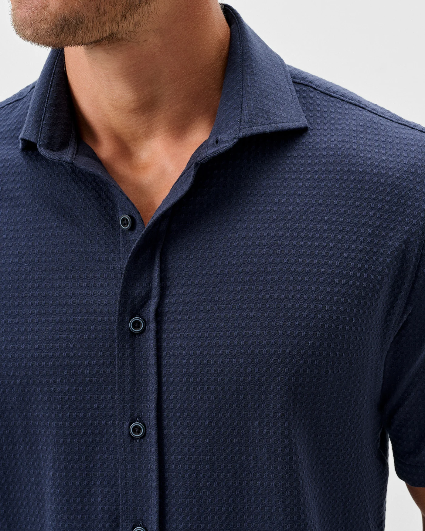 A man models the Johnnie-O Surfsucker Knit Short Sleeve Button Down Shirt in navy, featuring a collar and four visible buttons. The textured shirt by Johnnie-O is shown from shoulders to mid-chest.