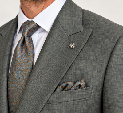 Close-up of a man in an olive Jack Victor Engel Olive Slim Fit Suit 3262602 with a white dress shirt, patterned paisley tie, matching pocket square, and metal lapel pin.