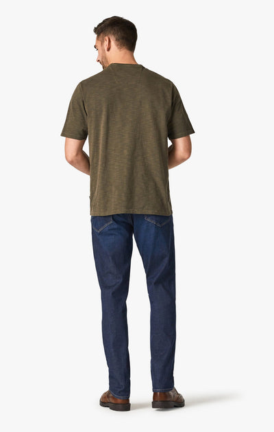 Man standing with his back to the camera, wearing a short-sleeve olive green shirt, 34 Heritage Dark Cashmere soft denim high rise straight leg jeans, and brown shoes against a plain white background.