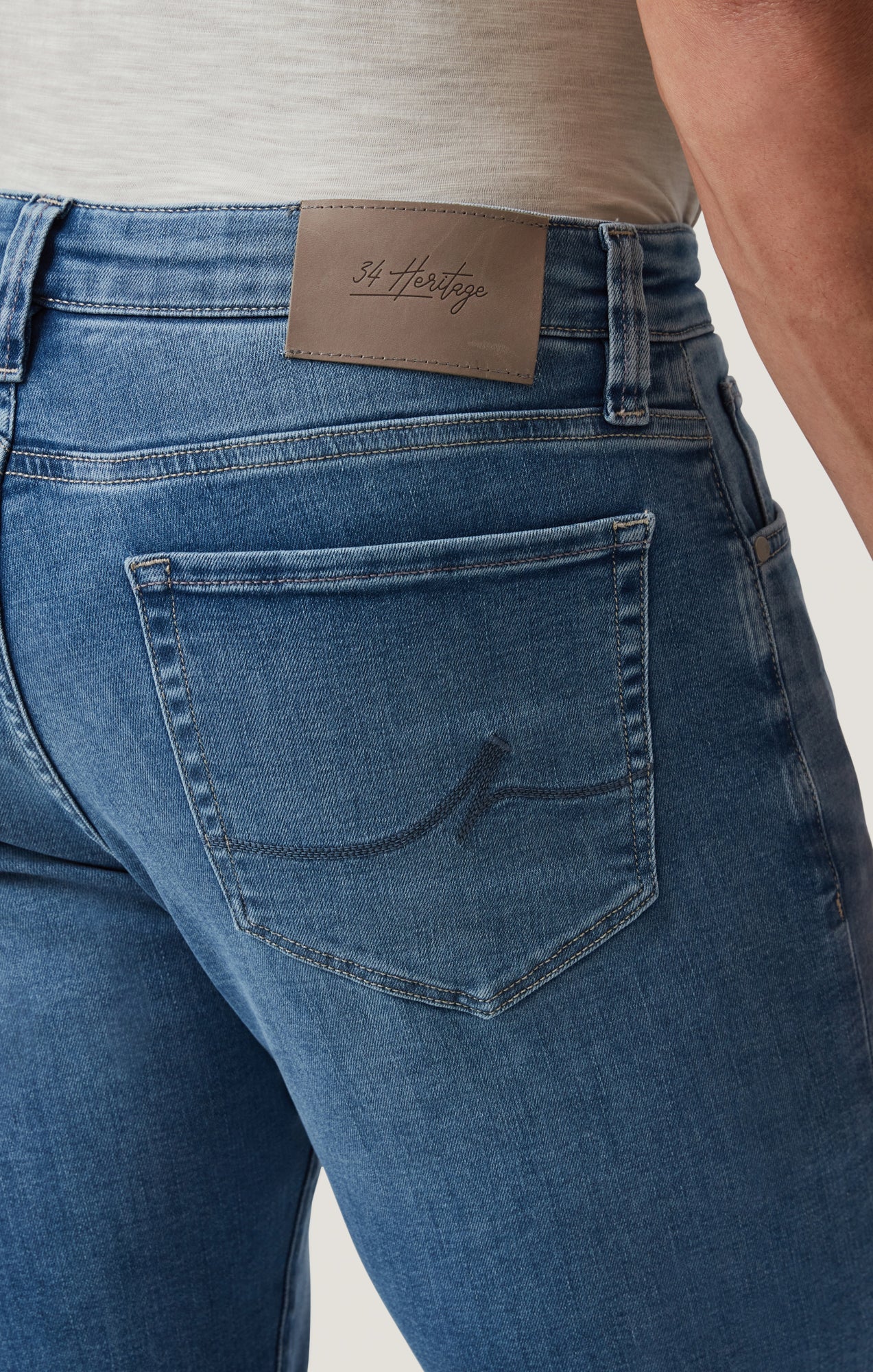 Close-up of a person wearing 34 Heritage Courage Jeans Organic Ultra in blue, paired with a light shirt, featuring a visible brown leather patch labeled 34 Heritage above the back pocket.