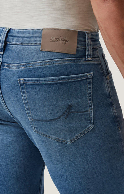 Close-up of a person wearing 34 Heritage Courage Jeans Organic Ultra in blue, paired with a light shirt, featuring a visible brown leather patch labeled 34 Heritage above the back pocket.