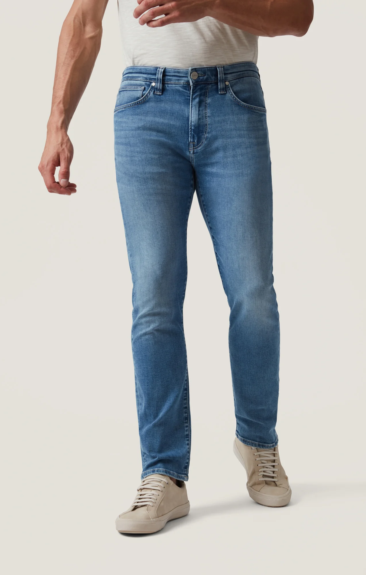 A person wearing 34 Heritage Courage Jeans Organic Ultra in light blue, paired with a white t-shirt and beige sneakers, stands against a plain background.