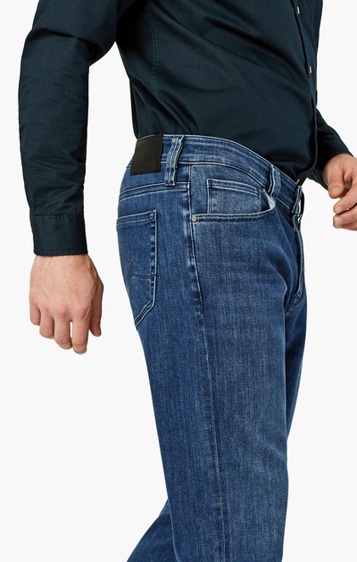 A person is shown from the waist down wearing 34 Heritage Charisma Mid Urban blue straight leg jeans made from premium stretch denim and a dark long-sleeve shirt, standing with their left arm relaxed by their side.
