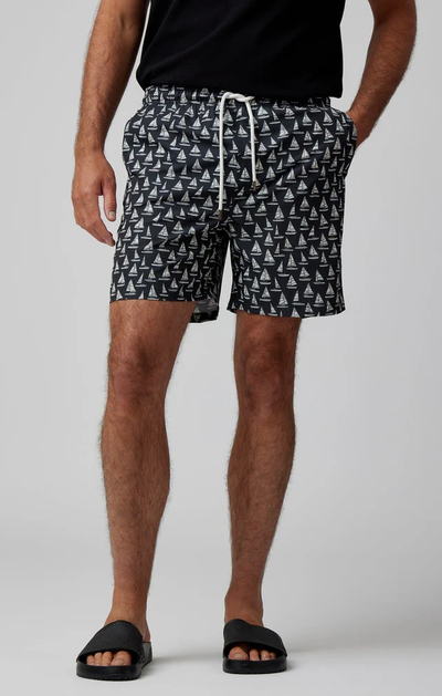 A person wearing a black t-shirt, Stone Rose HELIOS - Black Sailboats Swim Shorts, and black slides stands against a plain background, showcasing stylish men's swimwear by Stone Rose.