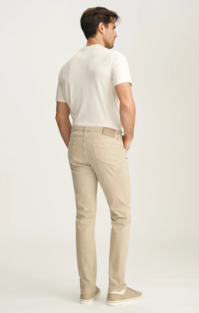 A man stands with his back to the camera, wearing a white t-shirt and 34 Heritage Courage Jeans Urban by 34 Heritage, paired with beige sneakers against a plain light background.