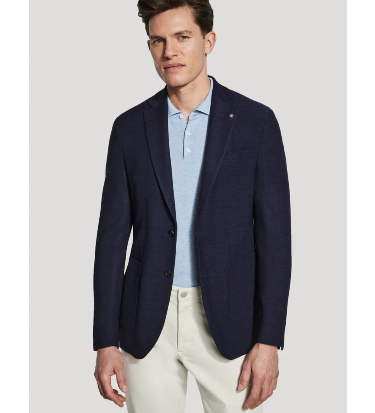 A man wearing the Jack Victor Hampton SC C1273 sport coat in a tailored fit, crafted from 100% wool by Jack Victor, layered over a light blue polo shirt and white pants, stands against a plain light gray background.