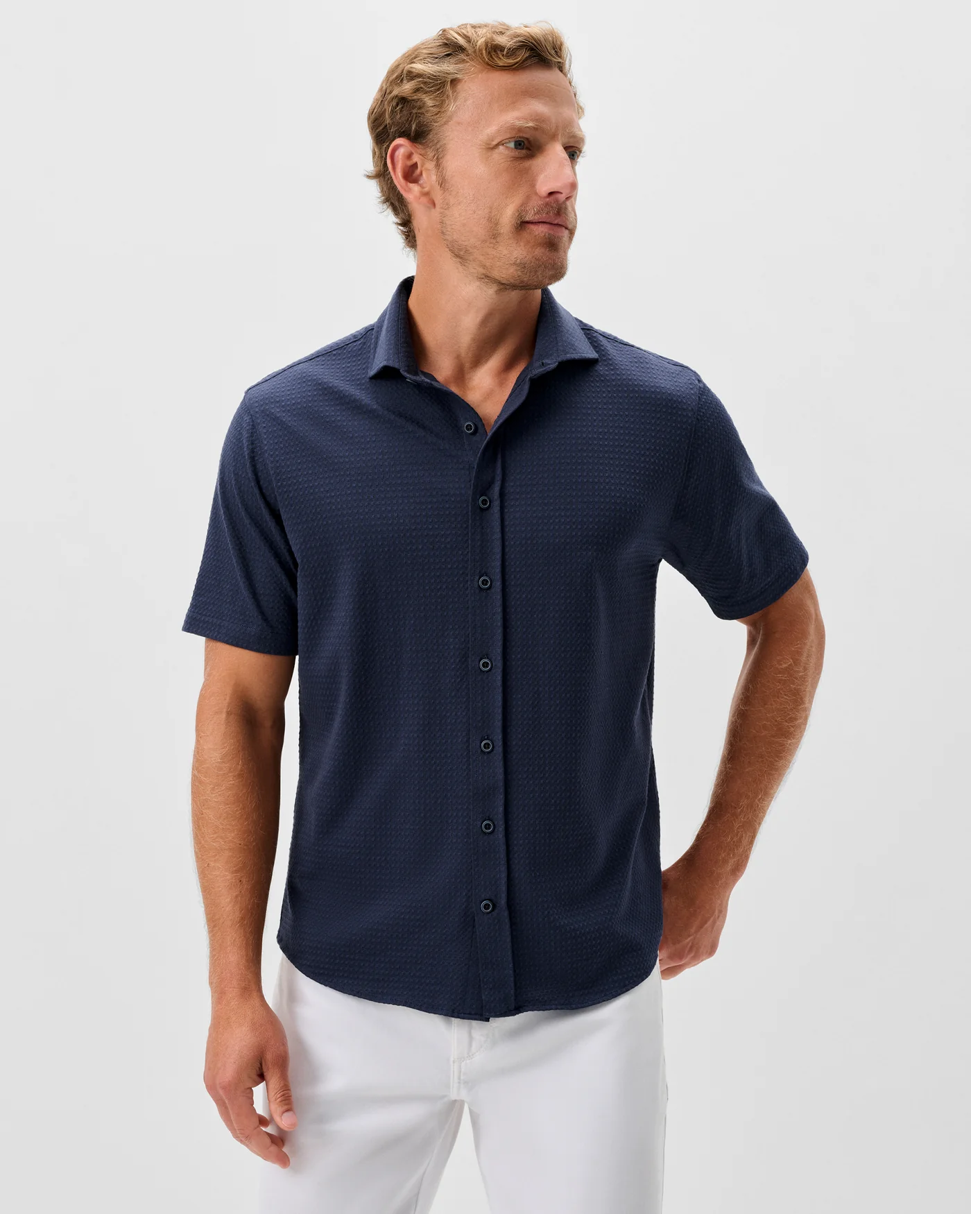 A man stands against a plain light background, facing slightly to the side, wearing the Johnnie-O Surfsucker Knit Short Sleeve Button Down Shirt in navy with white pants.