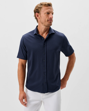 A man stands against a plain light background, facing slightly to the side, wearing the Johnnie-O Surfsucker Knit Short Sleeve Button Down Shirt in navy with white pants.