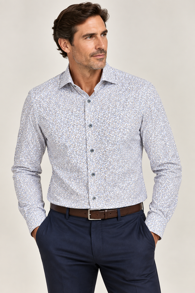 A man wearing the Elite Foliage Dress Shirt EX2674152 by Elite—a light patterned 100% cotton shirt—tucked into navy blue trousers with a brown belt, stands with his hands in his pockets against a plain background.
