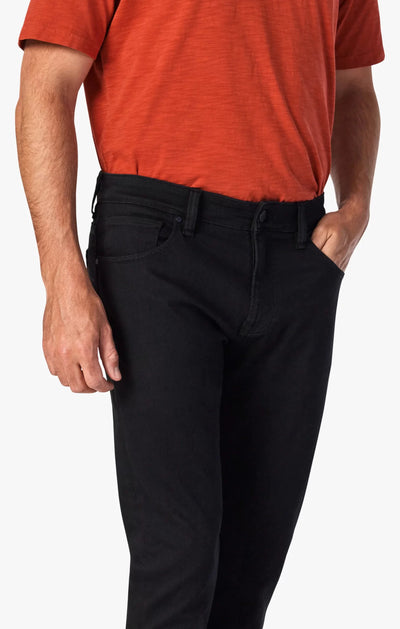 A person wearing a short-sleeved orange shirt and 34 Heritage Courage Black Urban Jeans stands with one hand in their pocket against a plain background.