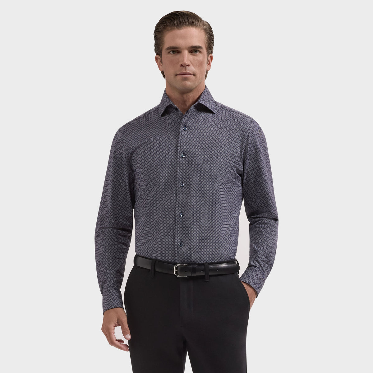 A man stands against a plain background, wearing the Bugatchi Devon Medallion Sport Shirt (KF9433K80), a dark geometric print button-up by Bugatchi, tucked into black pants with a belt.