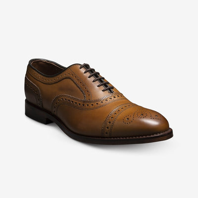 The Allen Edmonds Strand by Allen Edmonds is a brown calfskin oxford dress shoe with brogue detailing, black laces, and a low heel, shown against a white background.