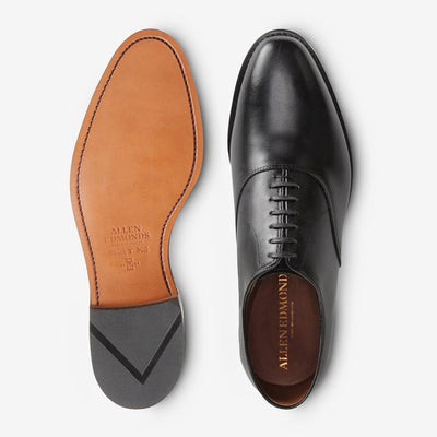The Allen Edmonds Carlyle by Allen Edmonds is a pair of black handcrafted leather oxford dress shoes, shown from the top and bottom, with black laces, a brown leather sole, and classic bench welt construction.