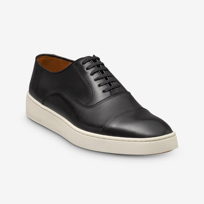 The Allen Edmonds Park Avenue Oxford Dress Sneaker by Allen Edmonds is a black premium leather shoe with a white rubber sole, cap toe, and black laces, displayed at an angle on a plain white background.