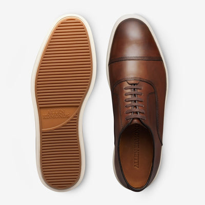 The Allen Edmonds Park Avenue Oxford Dress Sneaker features premium brown leather uppers, white rubber soles with Allen Edmonds branding, and is shown from the top and bottom.