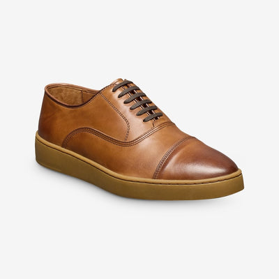 The Allen Edmonds Park Avenue Oxford Dress Sneaker features premium brown leather, a classic cap toe, lace-up closure, and rubber sole by Allen Edmonds, displayed on a plain white background.