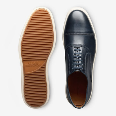 The Allen Edmonds Park Avenue Oxford Dress Sneaker features premium navy blue leather, white soles, and a tan textured rubber outsole—blending modern style with classic craftsmanship.