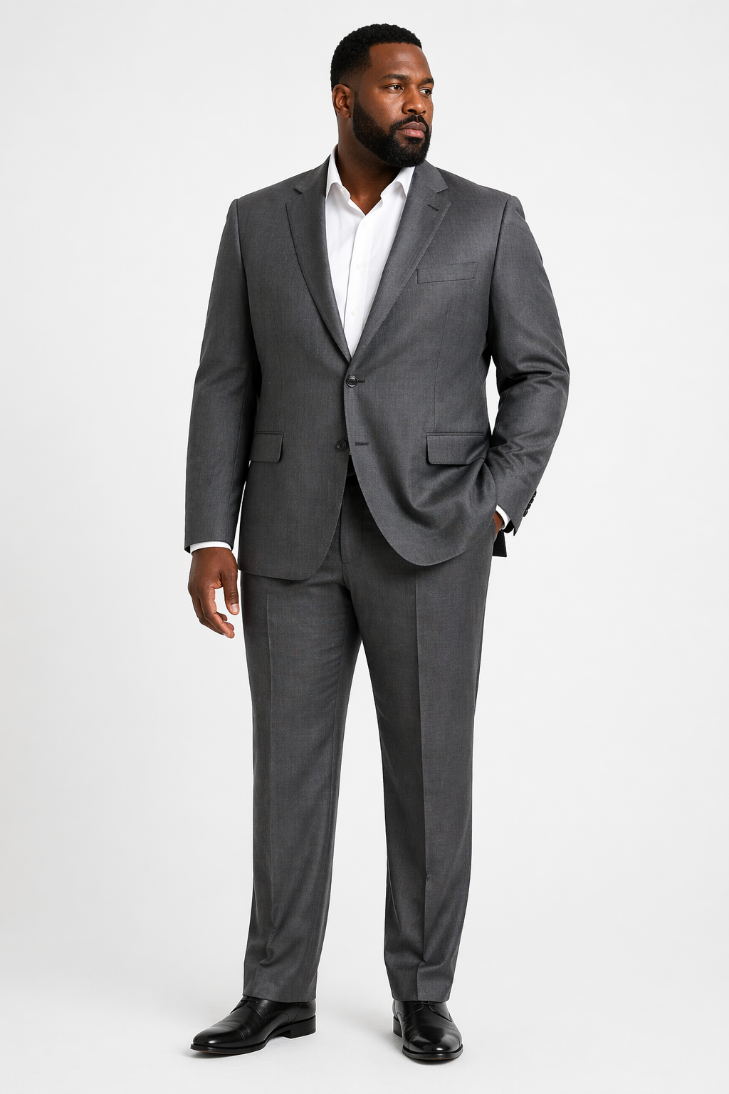 A man wears the Baroni Big & Tall Suit Notch Lapel in grey by Baroni, paired with a white dress shirt and black dress shoes, standing against a plain white background with one hand in his pocket.