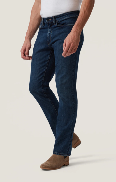 A person in 34 Heritage Charisma Mid Comfort jeans, a white shirt, and brown suede boots stands against a plain background.