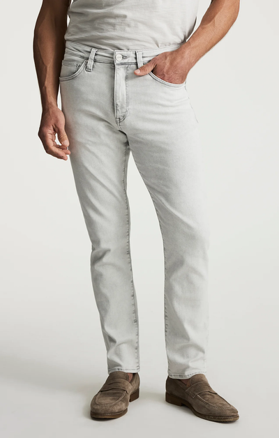 A person wearing 34 Heritage Courage Jeans Organic Ultra in light gray, paired with a white T-shirt and brown suede loafers, standing with one hand in a pocket.
