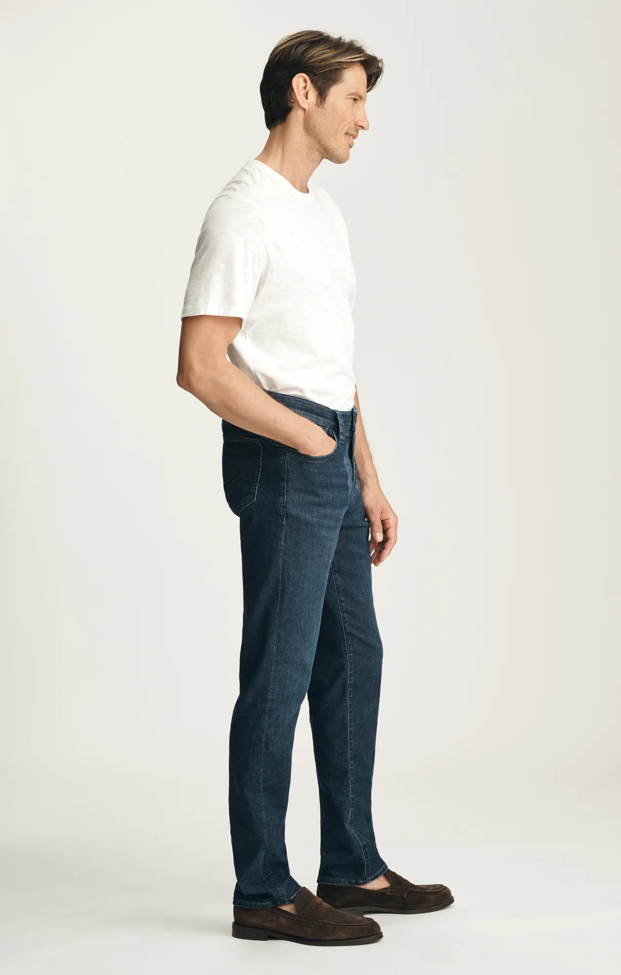 A man stands in profile against a plain background, wearing a white short-sleeve shirt and 34 Heritage Courage Sporty Jeans in dark blue denim featuring Thermocool fiber technology by 34 Heritage, paired with brown shoes and his hands in his pockets.