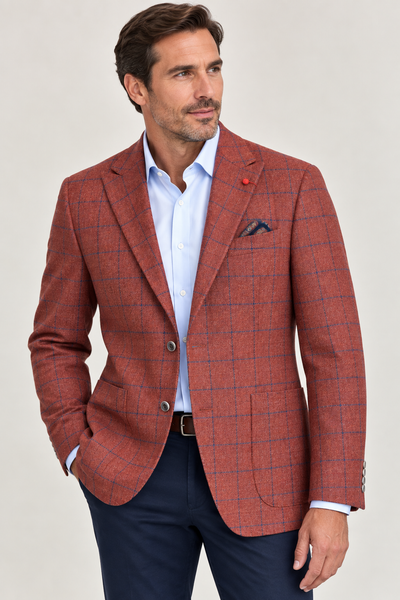 A man wearing the TailoRed Garro Rust Sport Coat 8110694—crafted from luxurious Italian lambswool cashmere by TailoRed—in a rich rust plaid, paired with a light blue dress shirt, navy pants, and a pocket square. He stands with one hand in his pocket and looks to the side against a neutral background, showcasing the perfect statement sport coat for men this fall.