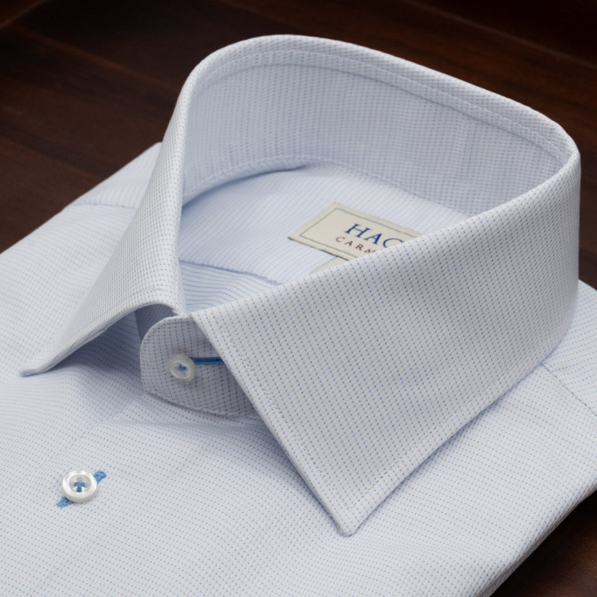 The Hagen Micro Dot Shirt MB7940100 by Hagen is a neatly folded light blue dress shirt with a spread collar, featuring a subtle micro dot jacquard pattern and white buttons. Crafted from 100% cotton for a modern fit, it is elegantly presented on a dark wooden surface.