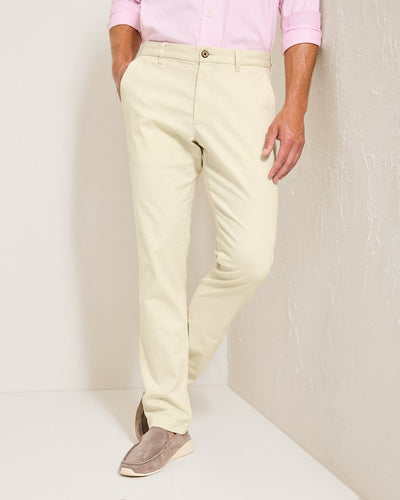 A person wearing Tommy Bahama Boracay Pant Big & Tall chinos from Tommy Bahama in light beige, paired with a light pink shirt with rolled-up sleeves and light brown loafers, stands against a white wall.