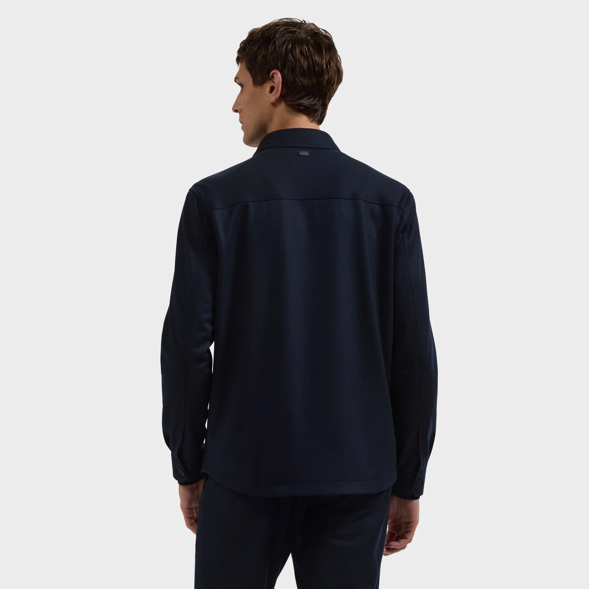 A man with short brown hair is standing and facing away, wearing a long-sleeve dark navy Bugatchi Button Shirt Jacket KX9015JS1 against a plain light gray background.