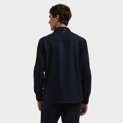 A man with short brown hair is standing and facing away, wearing a long-sleeve dark navy Bugatchi Button Shirt Jacket KX9015JS1 against a plain light gray background.