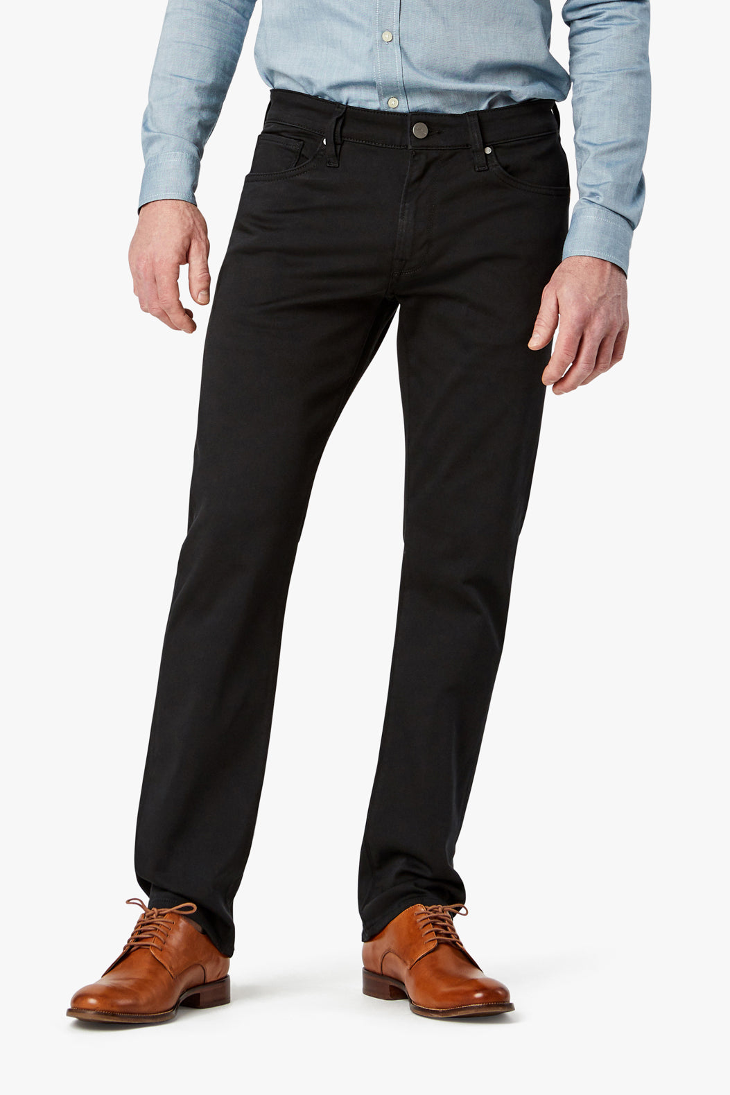 A man wearing a light blue button-up shirt, 34 Heritage Courage Double Black jeans by 34 Heritage, and brown dress shoes stands against a plain white background.