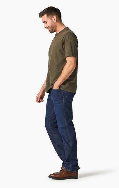 A man in a green t-shirt and brown boots stands in profile, smiling with one hand in his pocket, wearing 34 Heritage Dark Cashmere jeans by 34 Heritage against a plain white background.