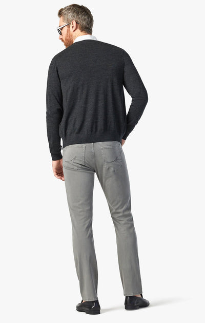 A man stands with his back to the camera against a white background, wearing a dark sweater, white collared shirt, black shoes, and 34 Heritage Pewter Twill relaxed straight-leg gray jeans.