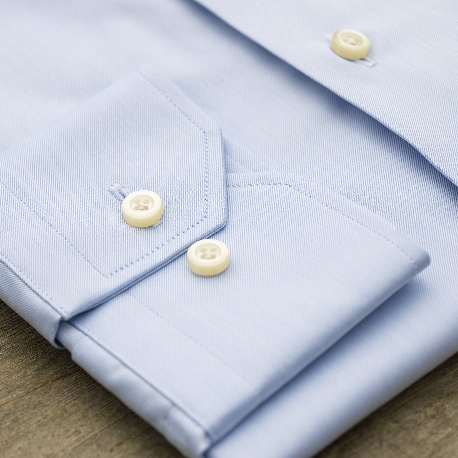 Close-up of the Hagen Solid Twill Shirt MB1864400 by Hagen, a neatly folded blue dress shirt crafted from 100% cotton superfine twill, highlighting the cuff and two white buttons on a wooden surface.