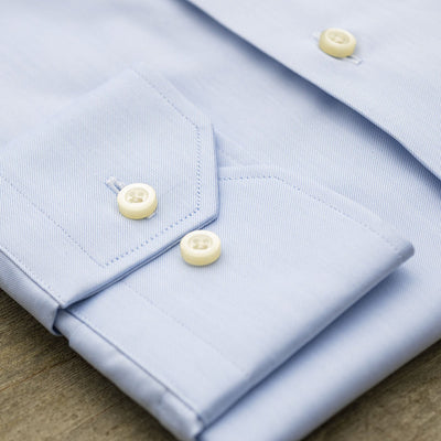 Close-up of the Hagen Solid Twill Shirt MB1864400 by Hagen, a neatly folded blue dress shirt crafted from 100% cotton superfine twill, highlighting the cuff and two white buttons on a wooden surface.