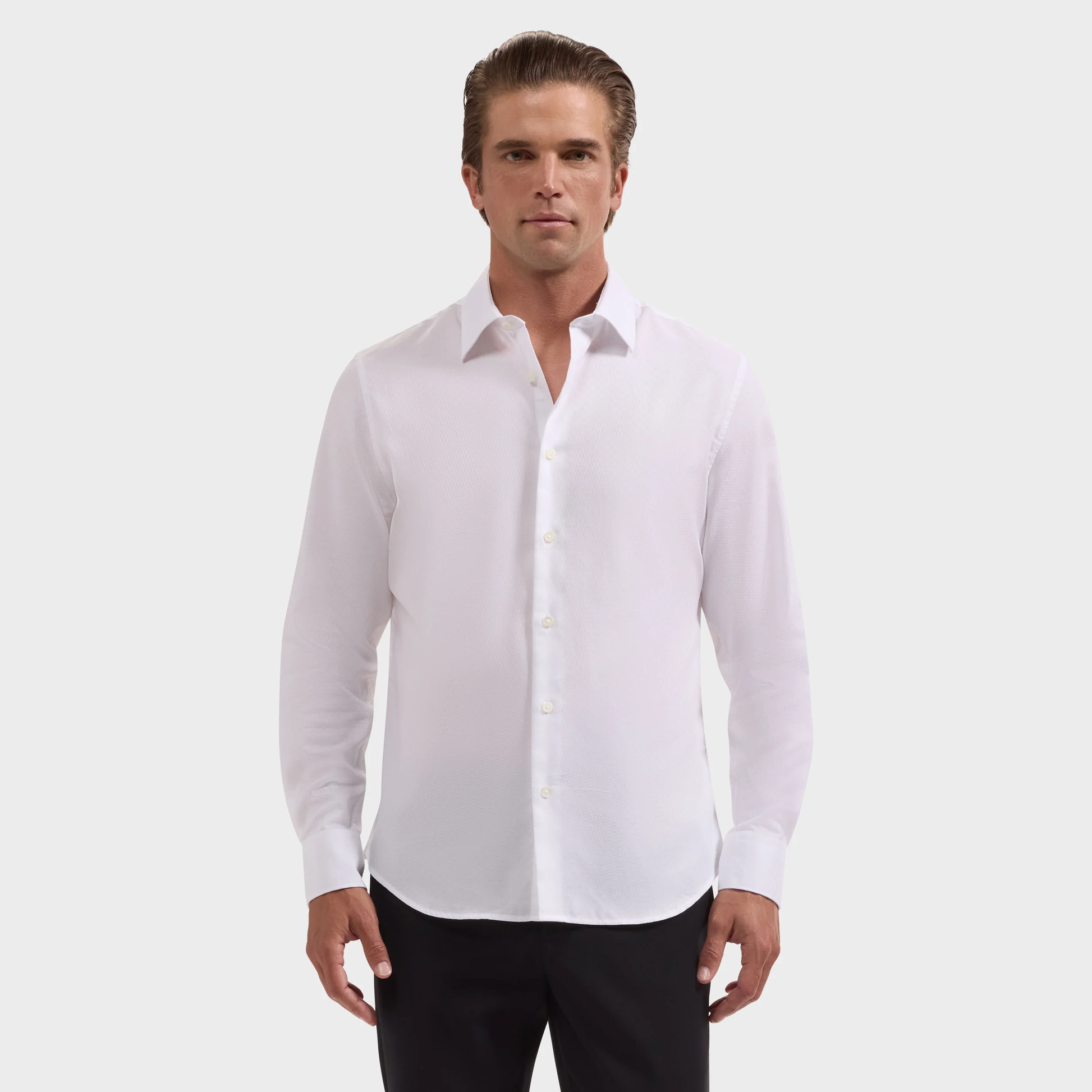 A man stands facing forward against a plain light background, wearing the Bugatchi Julian Tonal Geometric Jacquard Shirt KS4001L3S in white with long sleeves and black pants.