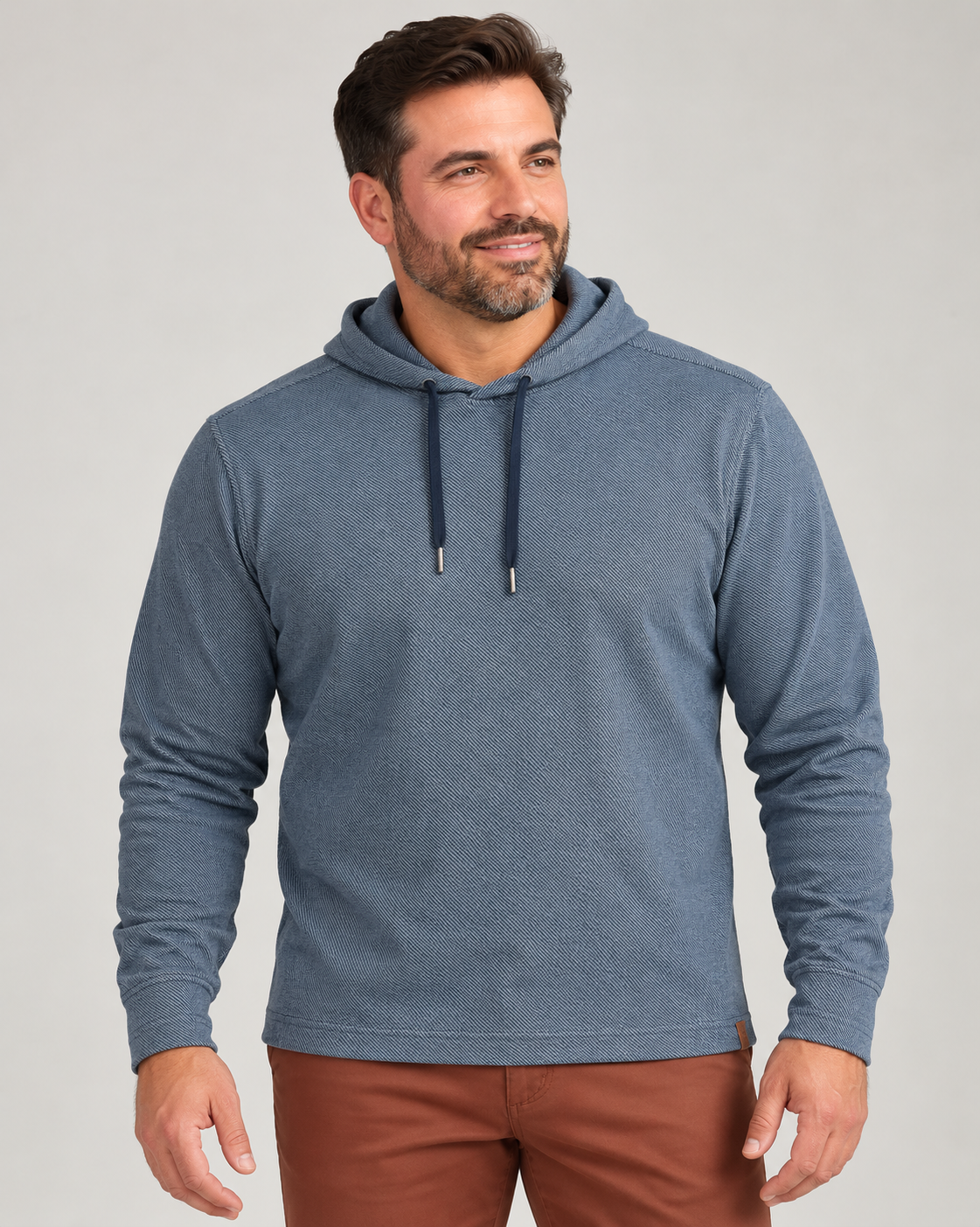 A man wearing a blue textured Tommy Bahama Fireside Coast Hoodie with a drawstring hood and brown trousers.