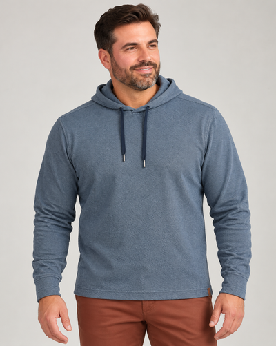 A man wearing a blue textured Tommy Bahama Fireside Coast Hoodie with a drawstring hood and brown trousers.