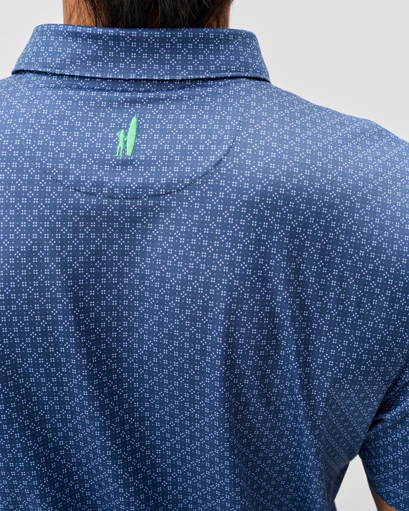 The back of a person wearing a Johnnie-O Torres Performance Big & Tall Polo featuring short sleeves, a blue pattern, and a small green surfboard-and-person logo below the collar.