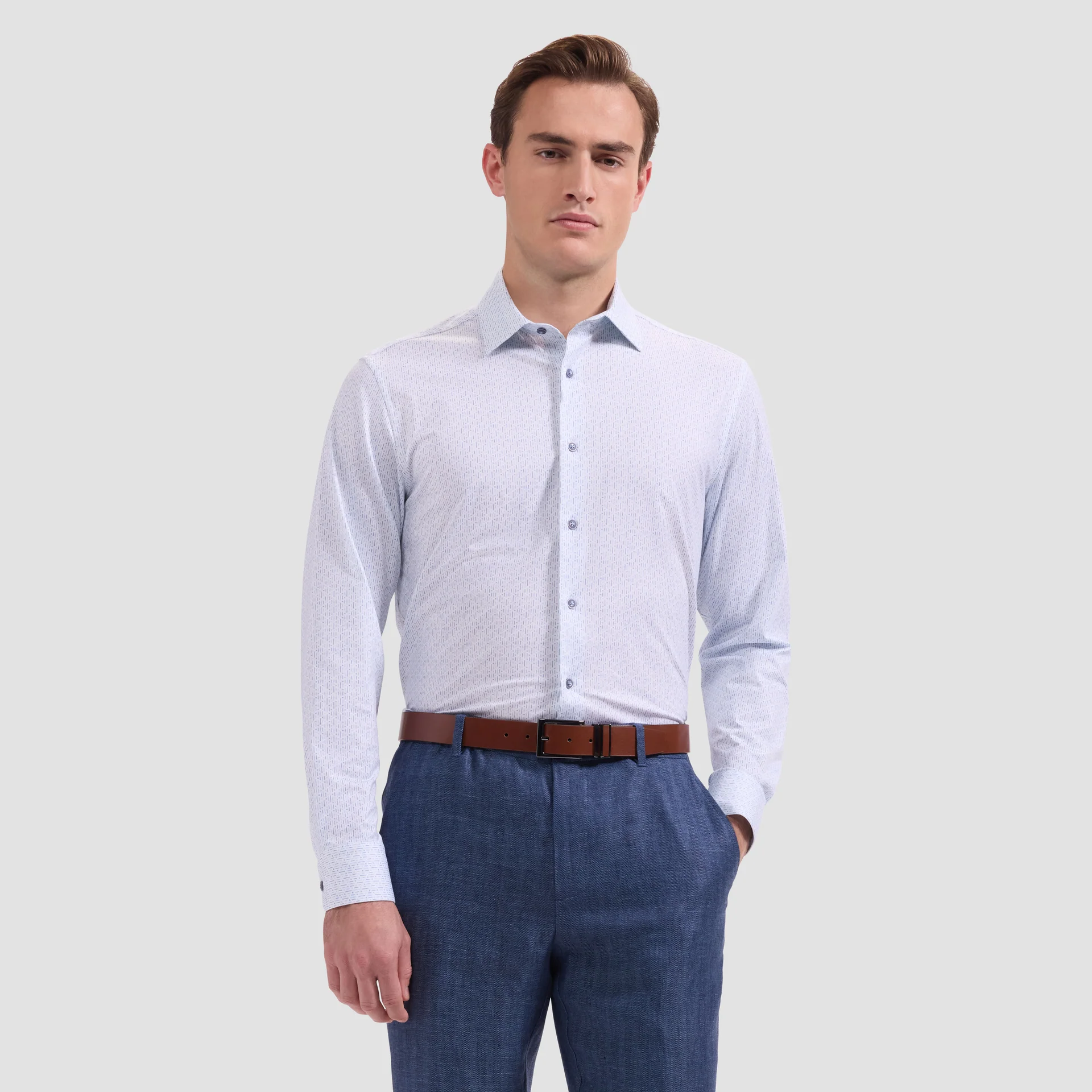 A man wearing the Bugatchi OP James OoohCotton® Shirt JF9216K60 with a geometric print, blue dress pants, and a brown belt stands with one hand in his pocket against a plain light background.
