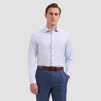 A man wearing the Bugatchi OP James OoohCotton® Shirt JF9216K60 with a geometric print, blue dress pants, and a brown belt stands with one hand in his pocket against a plain light background.