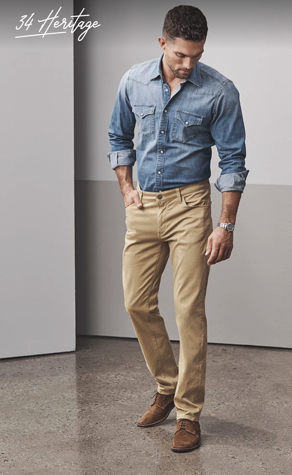 A man indoors wears 34 Heritage Charisma Khaki Twill chinos with a light denim shirt and brown shoes, head slightly down. "34 Heritage" is shown in the top left corner.