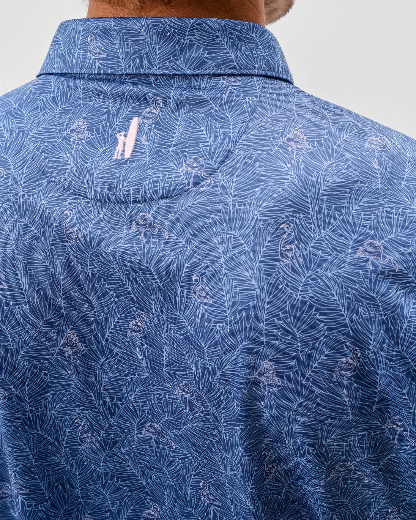 Close-up of the back of the Johnnie-O Osmond Big & Tall Polo by Johnnie-O, featuring a blue performance fabric with a white leaf and pink flamingo pattern, UPF 50 protection, moisture-wicking material, and a small pink embroidered logo near the collar.
