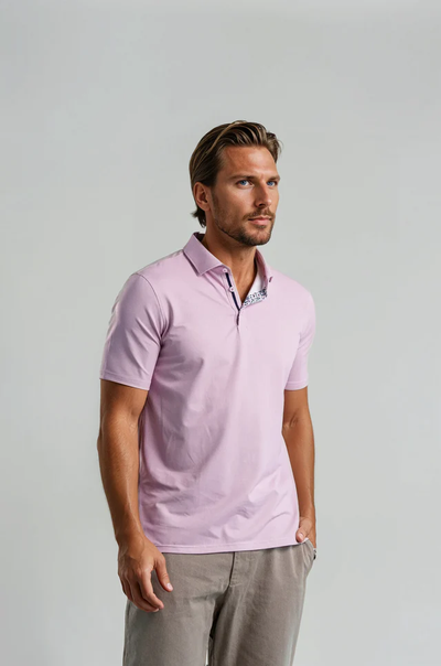 A man wears the Stone Rose Apex Birdseye Polo BSCSSP51 in light pink with gray pants, standing against a plain light gray background—creating a modern, classic summer look.