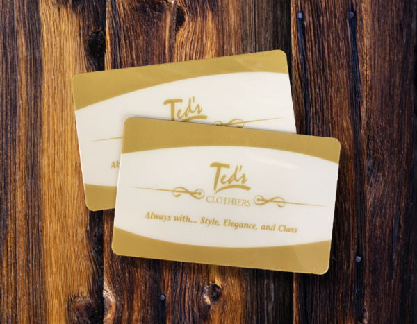 Two gold and cream-colored Gift Certificates from Ted's Clothiers, featuring the slogan "Always with... Style, Elegance, and Class," are displayed elegantly on a wooden surface.
