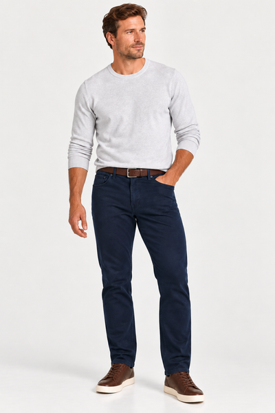 Man standing against a plain background, wearing a light gray sweater, Alberto PIPE 1607 Diamond Brushed Stretch Cotton Jeans in dark blue, a brown belt, and brown sneakers.