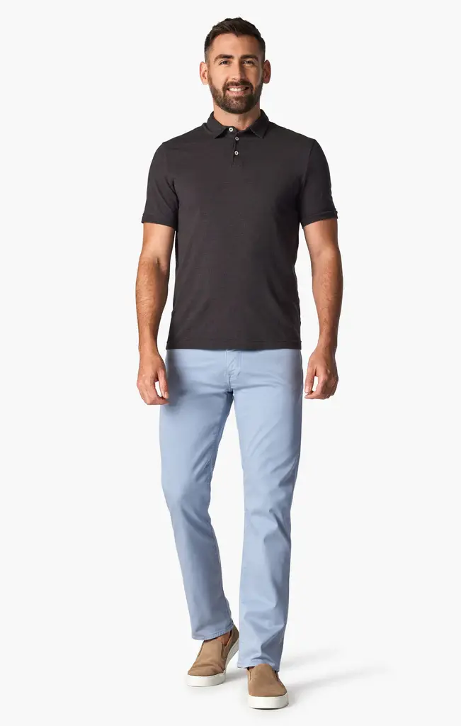 A man stands facing forward wearing a black polo shirt, tan slip-on shoes, and 34 Heritage Courage Coolmax modern straight leg light blue dress pants by 34 Heritage, against a plain white background.