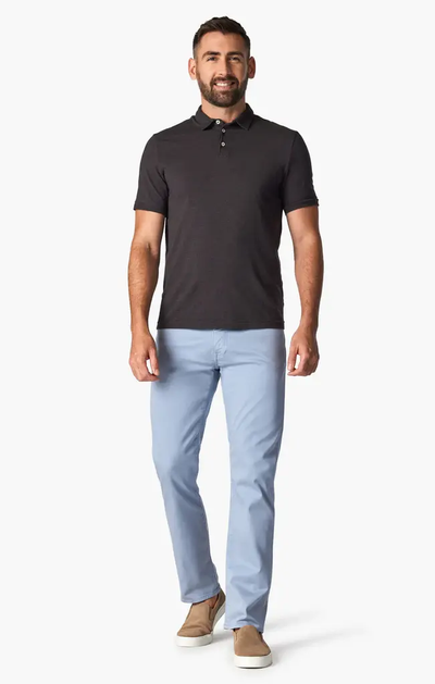 A man stands facing forward wearing a black polo shirt, tan slip-on shoes, and 34 Heritage Courage Coolmax modern straight leg light blue dress pants by 34 Heritage, against a plain white background.